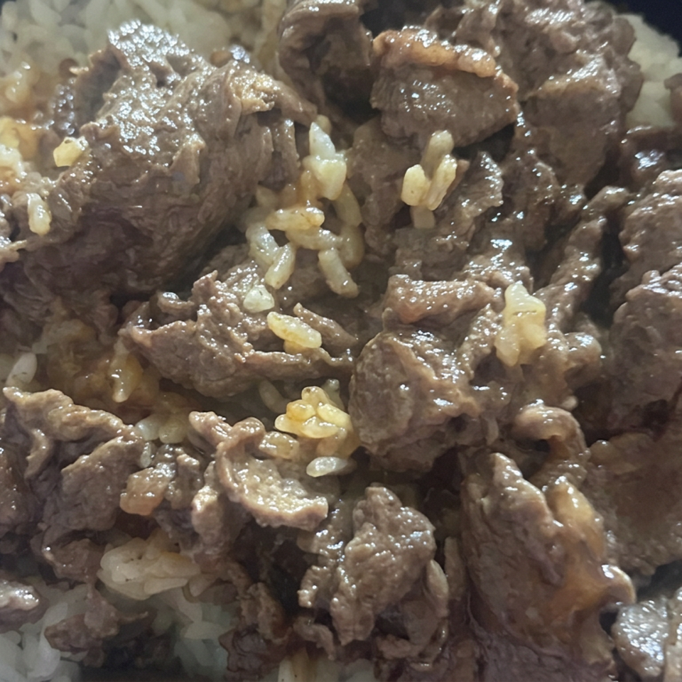 Close-up of saucy beef pieces served over white rice in a dark bowl.